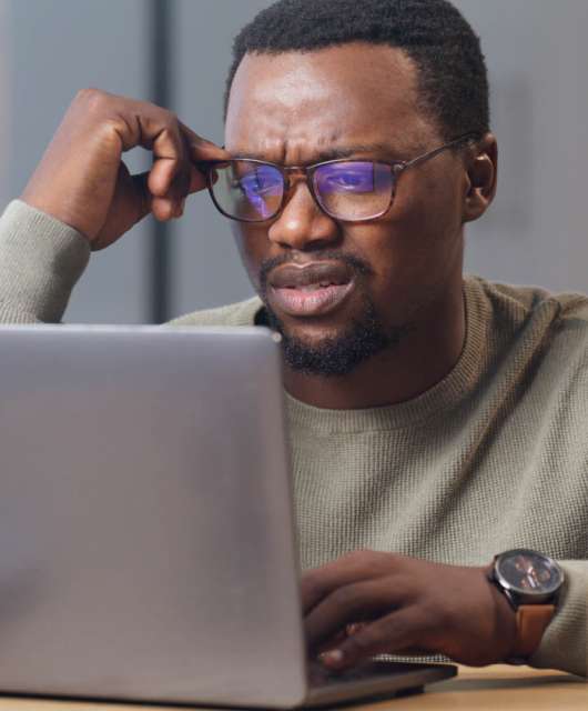 Man holding his glasses on his face while looking confused at a laptop screen.