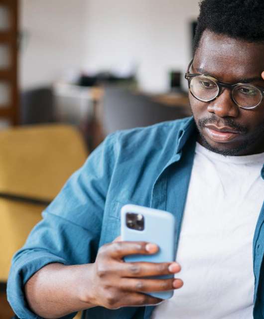 Black man in teal cardigan holding an iPhone with a blue case and looking confused.