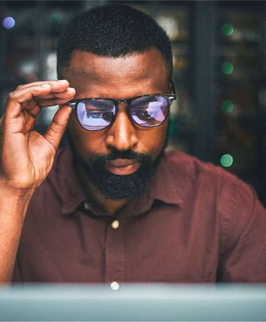 Black man holding his glasses while staring at a computer.