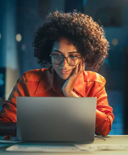 A woman sits at her laptop computer at night, looking intrigued